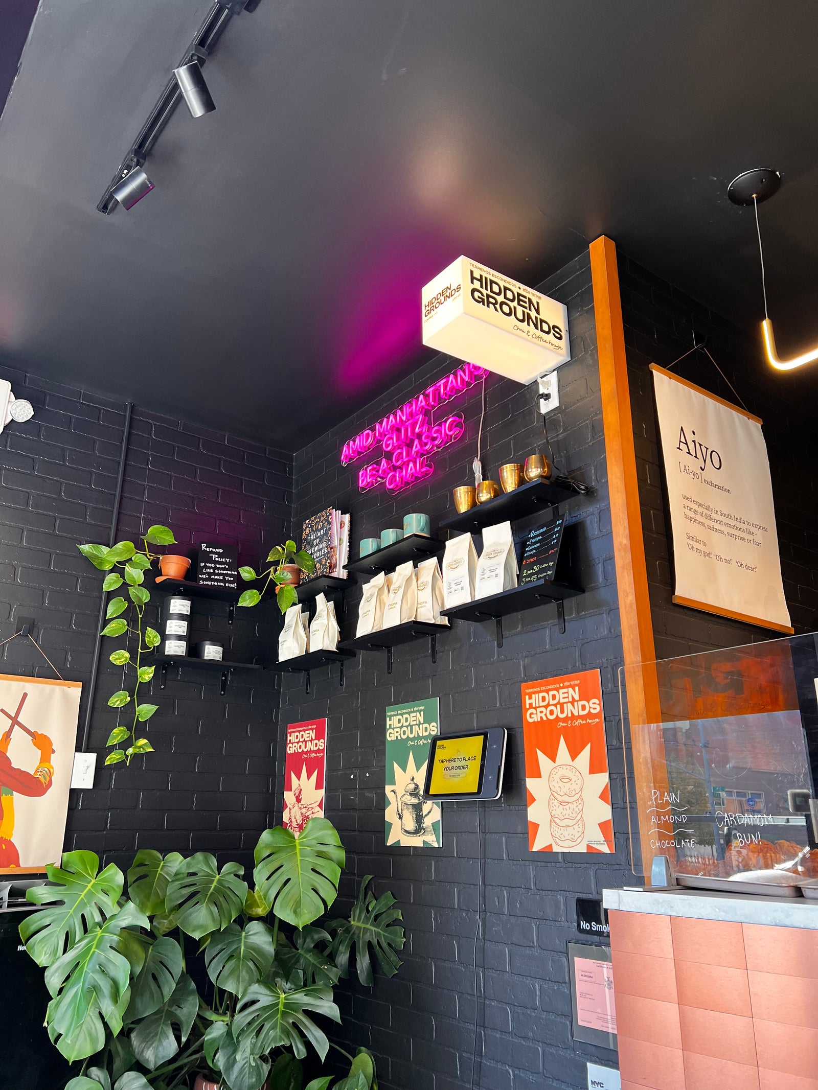 Black brick wall, coffee shelves, plants and a pink service counter at Hidden Grounds, 36 Third Avenue.